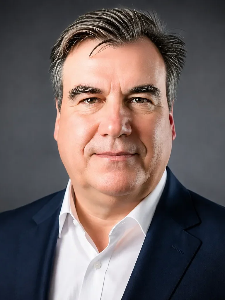 A man wearing a dark blue suit and white shirt against a dark background.